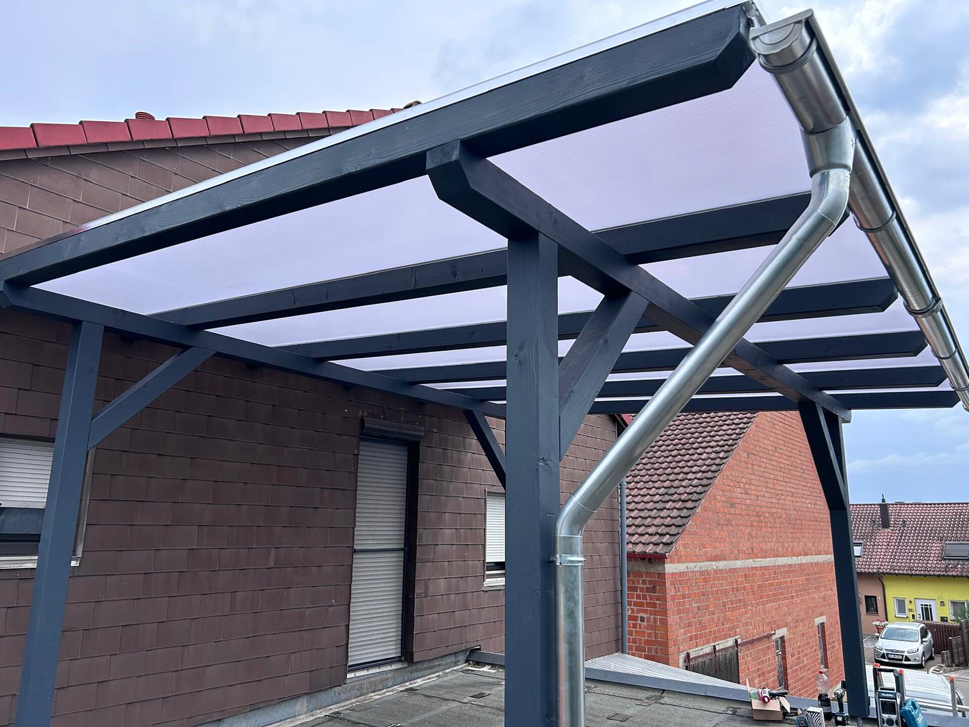 Carport mit anthrazitfarbenem Holzgerüst und lichtdurchlässigem Dach an einer Hauswand