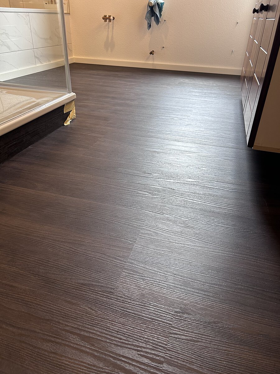 Dunkler Boden in Holzoptik in einem Badezimmer, im Hintergrund eine Duschkabine